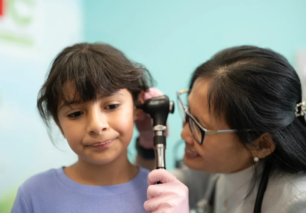 A healthcare professional examines a young girls ear using an otoscope. The girl, wearing a light blue shirt, looks slightly curious. The background is light blue with indistinct graphics.