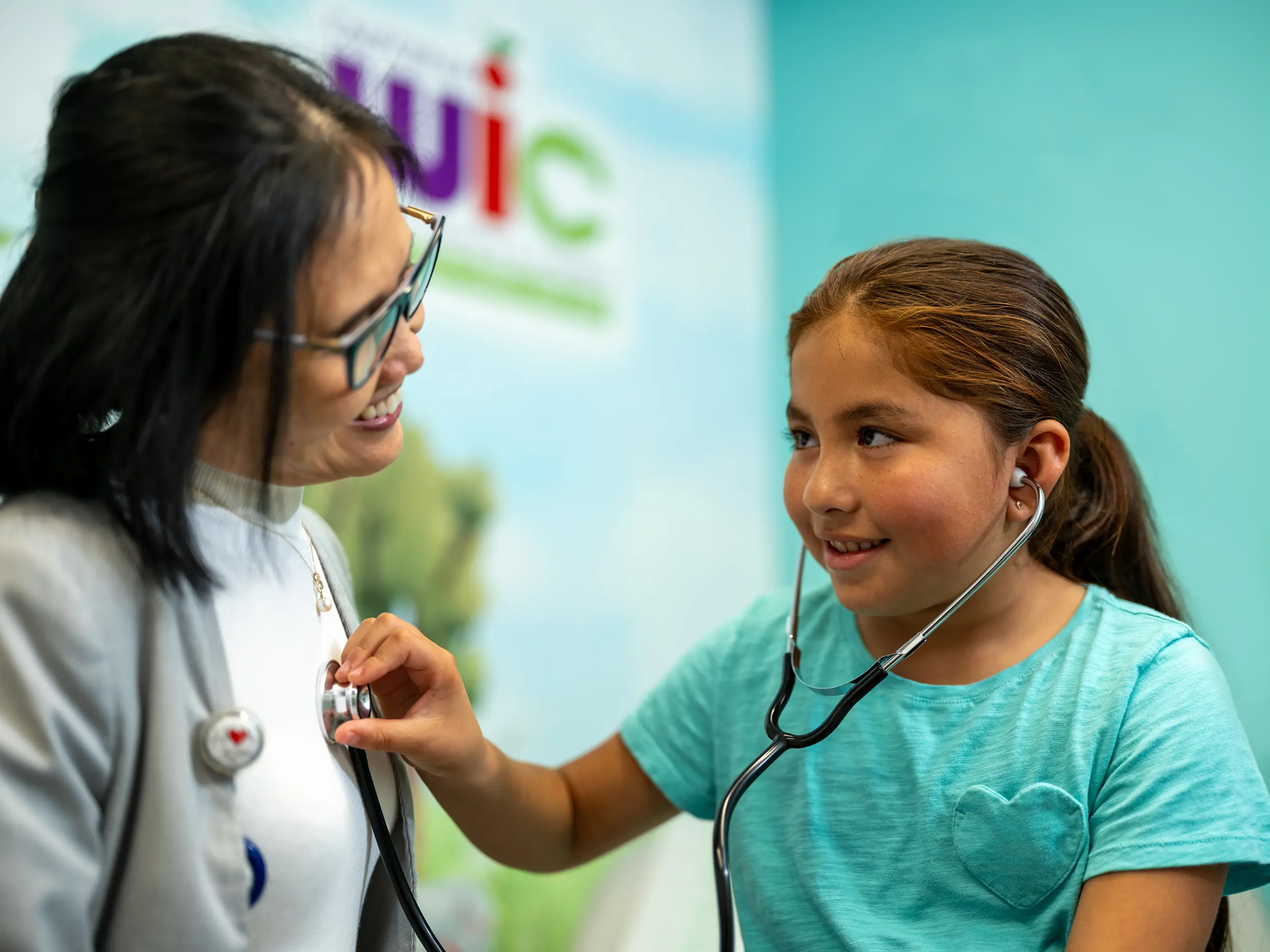 A girl wearing a teal shirt uses a stethoscope to listen to a womans heartbeat. The woman, wearing glasses and a gray jacket, smiles at the girl. They are in a brightly colored room with faint text in the background.