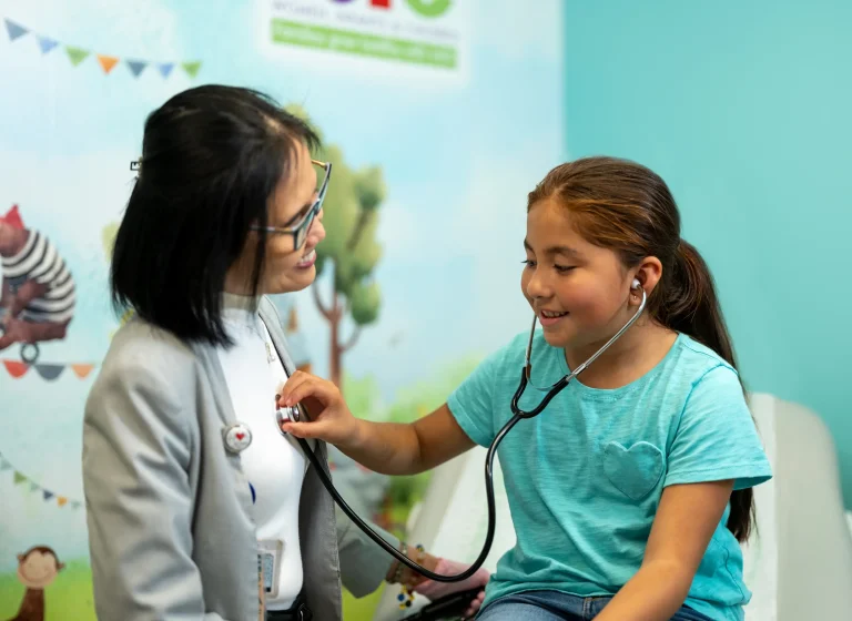 A young girl wearing a teal t-shirt listens to a female doctors heartbeat using a stethoscope in a bright medical office. The doctor smiles, engaging warmly. The room features colorful childrens decor.